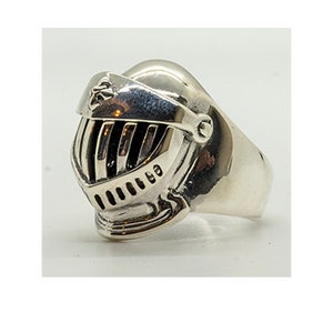 Puede incluir: Un anillo de plata con un diseño detallado de casco de caballero. El casco tiene una visera y una cresta en la parte superior.