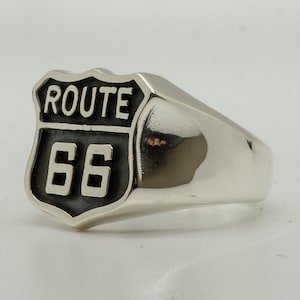 May include: A silver ring featuring the iconic Route 66 shield design. The shield is black with white lettering and the number 66. The ring has a polished, smooth band.