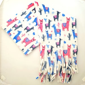 May include: A white fleece scarf featuring a repeating pattern of pink, blue, and gray llamas and cacti. The scarf has a fringed edge and is designed to be worn around the neck. The llama scarf is a fun accessory.