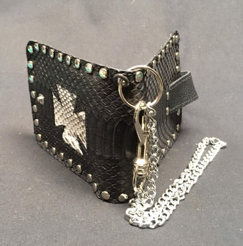 K&ouml;nnte beinhalten: Schwarze Ledergeldb&ouml;rse mit einer silbernen Kette und einem silbernen Verschluss. Die Geldb&ouml;rse hat ein Schlangenhautmuster und ist mit silbernen Nieten besetzt. Die Geldb&ouml;rse hat ein wei&szlig;es Lederpfeil-Design auf der Vorderseite.