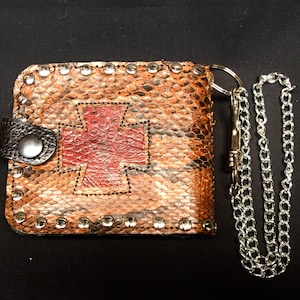 Può includere: Un portafoglio in pelle marrone con un design a croce rossa e una catena argentata. Il portafoglio ha un bordo borchiato e una chiusura a lembo.
