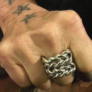 May include: A silver ring with a woven chain design. The ring is worn on a hand with star tattoos. The ring's intricate design is made of multiple interwoven links, creating a bold and textured appearance. The ring is a statement piece.