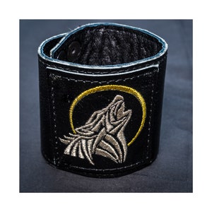 May include: Black leather wrist cuff with a silver embroidered howling wolf design inside a gold circle. The cuff has a snap closure and is lined with black leather. A stylish accessory.
