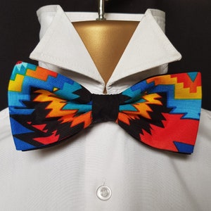 May include: A colorful bow tie with a geometric pattern in shades of red, orange, yellow, blue, and black. The bow tie is made of fabric and has a black adjustable strap.