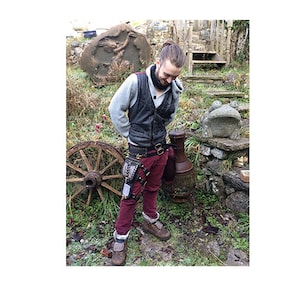 Könnte beinhalten: Eine Person trägt eine schwarze Lederweste, einen grauen Pullover und weinrote Hosen. Ein schwarzer Gürtel mit einer Tasche und Riemen ist um die Taille. Die Person trägt braune Stiefel. Ein altes Holzrad und eine steinerne Froschstatue befinden sich im Hintergrund.