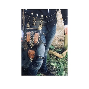 Könnte beinhalten: Ein schwarzes Leder-Steampunk-Oberschenkelholster mit einer braunen Ledertasche und Metallverzierungen. Das Holster wird über einer blauen Jeans und einem schwarzen Gürtel mit Metallnieten getragen.