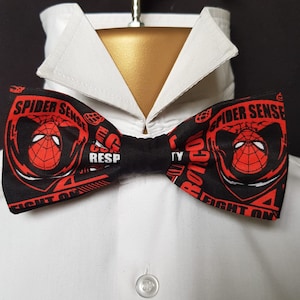 Puede incluir: Pajarita negra con un diseño de Spider-Man rojo y blanco. La pajarita tiene el texto "Spider Sense" y "Fight On".