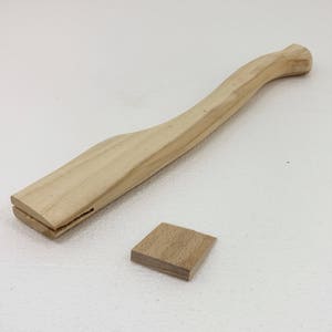 Op de afbeelding: Een houten bijl steel met een gebogen vorm en een klein houten wigje. De steel is onbehandeld en heeft een natuurlijke houtkleur.