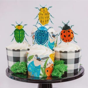 Può includere: Cupcake con glassa bianca e decorazioni a tema insetti. I cupcake sono in pirottini a scacchi bianchi e neri e fantasia. I topper presentano un design a forma di coleottero giallo, verde, blu e arancione. I cupcake sono su un'alzatina nera.