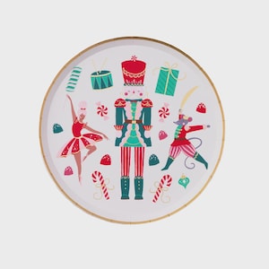 Puede incluir: Un plato redondo blanco con borde dorado con un tema festivo de Cascanueces. El diseño incluye un soldado cascanueces, una bailarina, un ratón, bastones de caramelo y otras ilustraciones temáticas navideñas en rojo, verde y azul.