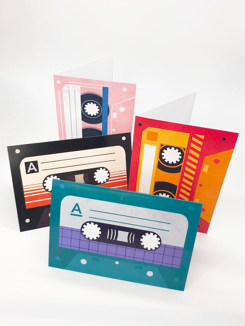 May include: Four colorful blank greeting cards with a cassette tape design. The cards are pink, red, black, and teal. Each card has a different design of a cassette tape with the letter "A" on the top.