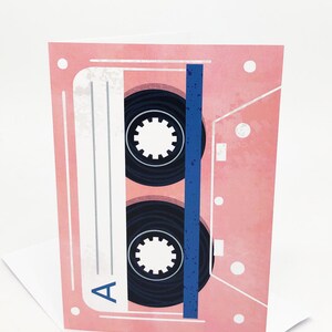 May include: A pink cassette tape with a blue stripe and black spools. The tape is labeled with the letter "A".
