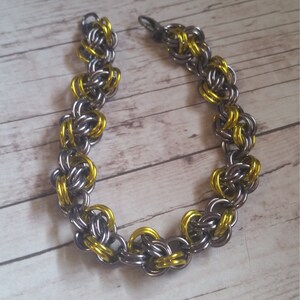 May include: A silver and gold chainmail bracelet with a clasp closure. The bracelet is made of small metal rings that are interlocked to create a chainmail pattern.