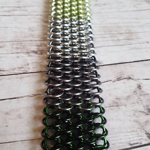 May include: A green, silver, and black chainmail bracelet. The bracelet is made of small metal rings that are linked together.