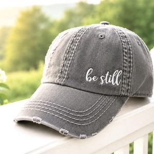 May include: A charcoal gray baseball cap with a distressed, worn-in finish. The front panel features the white embroidered text 'be still' in a cursive script font. The cap has a curved brim with visible stitching and frayed edges throughout.