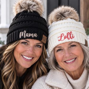May include: A black knit beanie with a faux fur pom-pom and the word Mama embroidered in white, alongside a white knit beanie with a faux fur pom-pom and the word Lolli embroidered in red. Both beanies feature a small brown C.C. brand tag.