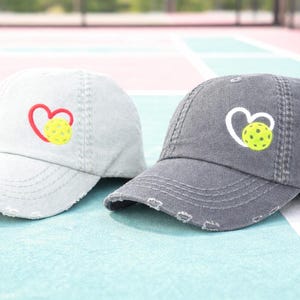 May include: Two distressed baseball caps, one light gray and one dark gray. Each cap features a heart-shaped design with a pickleball in the center. The caps have a worn, vintage look.