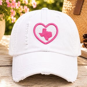 May include: White baseball cap with a distressed brim, featuring a pink heart outline with the state of Texas embroidered inside. The cap is on a wooden surface, with a blurred background of flowers and a straw bag.