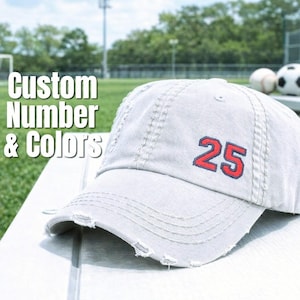 May include: A light gray distressed baseball cap with the number "25" embroidered in red. The cap has a curved brim and is sitting on a white surface. The text "Custom Number & Colors" is in the background.