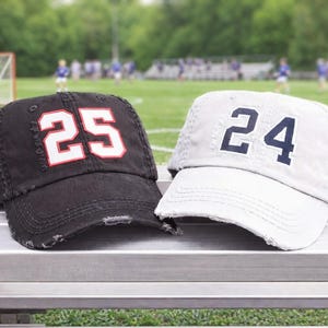May include: Two distressed baseball caps, one black with the number 25 in white and red, and one gray with the number 24 in navy blue. The caps are on a metal bench with a blurred sports field in the background.
