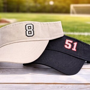 May include: Two visors, one tan and one black, rest on a wooden surface. The tan visor has the number 8 embroidered in black. The black visor has the number 51 embroidered in red. The background is a blurred green field.