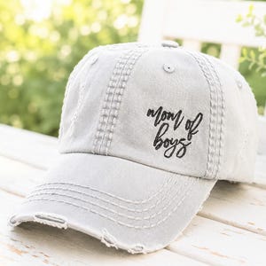 May include: A light gray distressed baseball cap featuring the embroidered black text "mom of boys" on the front panel. The cap has a curved brim with frayed edges, stitched detailing, and a structured crown with ventilation eyelets.