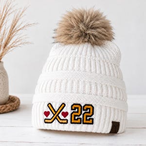 Op de afbeelding: Een witte, geribbelde gebreide muts met een grote bruine pompon van imitatiebont aan de bovenkant. De voorkant is voorzien van een geborduurde patch met twee gekruiste gele hockeysticks, twee kleine rode hartjes en het gele nummer 22 met een zwarte omlijning.