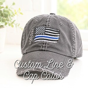 May include: A distressed gray baseball cap featuring an embroidered American flag design with a blue line. The cap has a pre-curved bill and adjustable strap. The text "Custom Line & Cap Color" is written on the front.