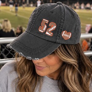 May include: A charcoal gray baseball cap with a distressed brim, featuring the number 52 embroidered in brown and white, alongside a small heart-shaped patch containing a white football graphic on the side of the crown.