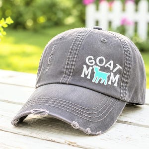 May include: A charcoal gray distressed baseball cap featuring the white text GOAT MOM with a light blue goat silhouette replacing the letter O. The cap has a curved brim with frayed edges and visible stitching details across the front panels.