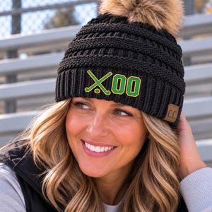 May include: A black knit beanie with a large brown faux fur pom-pom on top. The cuff features an embroidered graphic of two crossed hockey sticks and the number 00 in green with yellow outlines. A small brown leather tag with C.C. is attached.
