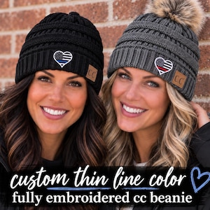 May include: Two cable-knit beanies with a faux fur pom-pom on the gray version. Both feature a heart-shaped patch with a thin line flag design. The black beanie has a blue line, and the gray beanie has a red line. Each has a brown C.C brand tag.