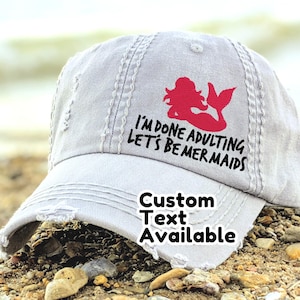 Op de afbeelding: Een grijze, verweerde baseballcap met de tekst "I'M DONE ADULTING LET'S BE MERMAIDS" en een rode zeemeermingrafiek. De cap heeft ook de tekst "Custom Text Available".