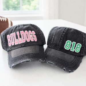 May include: Two distressed black baseball caps. One cap features the word "BULLDOGS" in pink lettering, and the other displays the number "818" in green. A brown football is in the background.