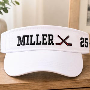 May include: White visor with the name "MILLER" in black block letters, crossed hockey sticks, and the number "25". The visor is a sports accessory, likely for sun protection.