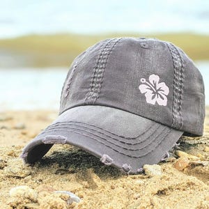 May include: A distressed gray baseball cap with a light pink hibiscus flower design. The cap has a curved brim and is sitting on a sandy surface. The cap has a worn, vintage look.