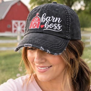 May include: A charcoal grey distressed baseball cap featuring an embroidered red barn icon, a small red heart, and the white cursive text "barn boss" centered on the front panel. The cap has a curved brim with frayed edges and a structured crown.