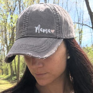 May include: A charcoal gray baseball cap with a distressed, worn-in finish. The front panel features the white script text "Mama" followed by a small, light pink heart icon. The cap has a curved brim with frayed edges and a button on top.