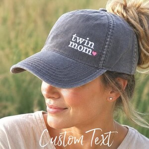 May include: A charcoal gray baseball cap featuring the white embroidered text twin mom with a small pink heart icon. The cap has a distressed, washed-out appearance and includes a high opening at the back for a ponytail or bun hairstyle.