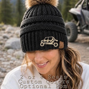 May include: A black knit beanie with a ribbed texture and a brown faux fur pom-pom on top. A tan leather patch on the front features a gold-colored graphic of a side-by-side off-road vehicle. The text 2 or 4 Seats Custom Colors Optional Pom is visible.
