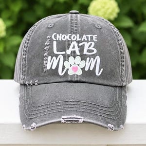Pu&ograve; includere: Un berretto da baseball grigio antracite dal look vissuto, con la scritta bianca "CHOCOLATE LAB MOM" e una grafica a forma di zampa bianca con un piccolo cuore rosa al centro. Il berretto presenta una finitura usurata con bordi sfilacciati.