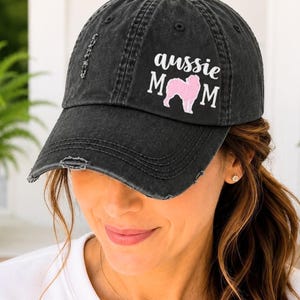 May include: A black distressed baseball cap featuring the white text "aussie" above the word "mom" in white, with a pink silhouette of an Australian Shepherd dog positioned between the letters. The cap has a worn, vintage-style finish.