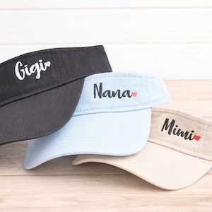 May include: Three visors in black, light blue, and beige. Each visor has a name embroidered in a script font: "Gigi", "Nana", and "Mimi". A small red heart is next to each name. The visors are on a light wood surface.