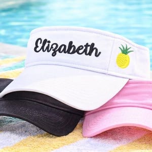 May include: Three visors in black, white, and pink rest on a striped towel near a pool. The white visor has the name "Elizabeth" embroidered in black script and a yellow pineapple graphic. The other visors are solid colors.