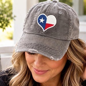 May include: A charcoal gray distressed baseball cap featuring a heart-shaped patch on the front. The patch displays the Texas state flag design, consisting of a vertical blue stripe with a white star, followed by horizontal white and red stripes.