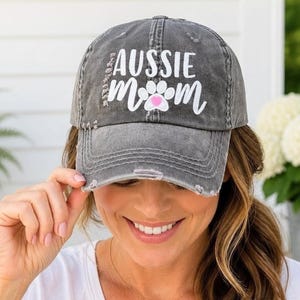 May include: A charcoal gray distressed baseball cap featuring the white text "AUSSIE MOM" with a white paw print graphic containing a small pink heart in the center. The cap has a worn, vintage-style finish with frayed edges on the brim and crown.