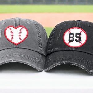 May include: Two baseball caps. One is gray with a heart-shaped baseball design. The other is black with a baseball design and the number 85. Both caps have a distressed, worn look.