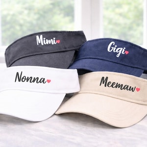 May include: Four visors in navy, black, white, and tan. Each visor has a name in script with a small red heart. The names are Mimi, Gigi, Nonna, and Meemaw. The visors are displayed in front of a window.