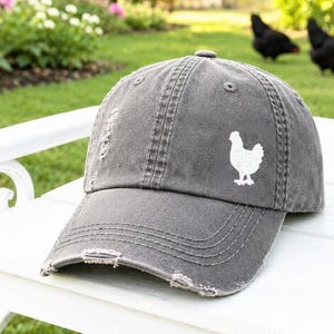May include: A charcoal gray baseball cap with a distressed, worn-in finish featuring a small, white embroidered chicken silhouette on the front panel. The cap has a curved brim with frayed edges and stitched eyelets for ventilation.
