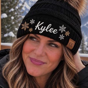 May include: A black knit beanie with a cable-knit texture, featuring a large brown faux fur pom-pom on top. The cuff displays the name Kylee in white script, surrounded by small gold and white snowflake icons. A small tan CC brand tag is attached.
